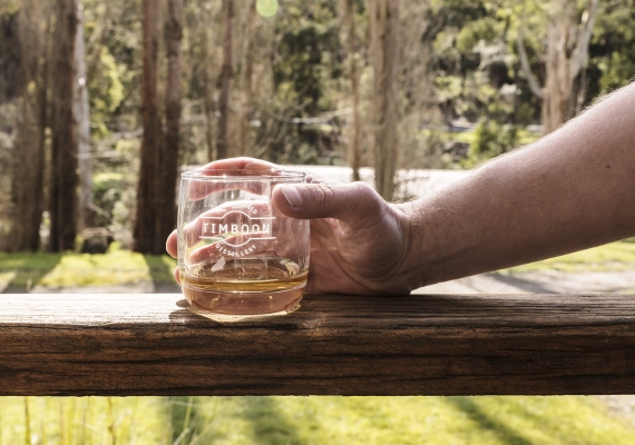 Hand holding a glass of whisky on a railing with trees in the background